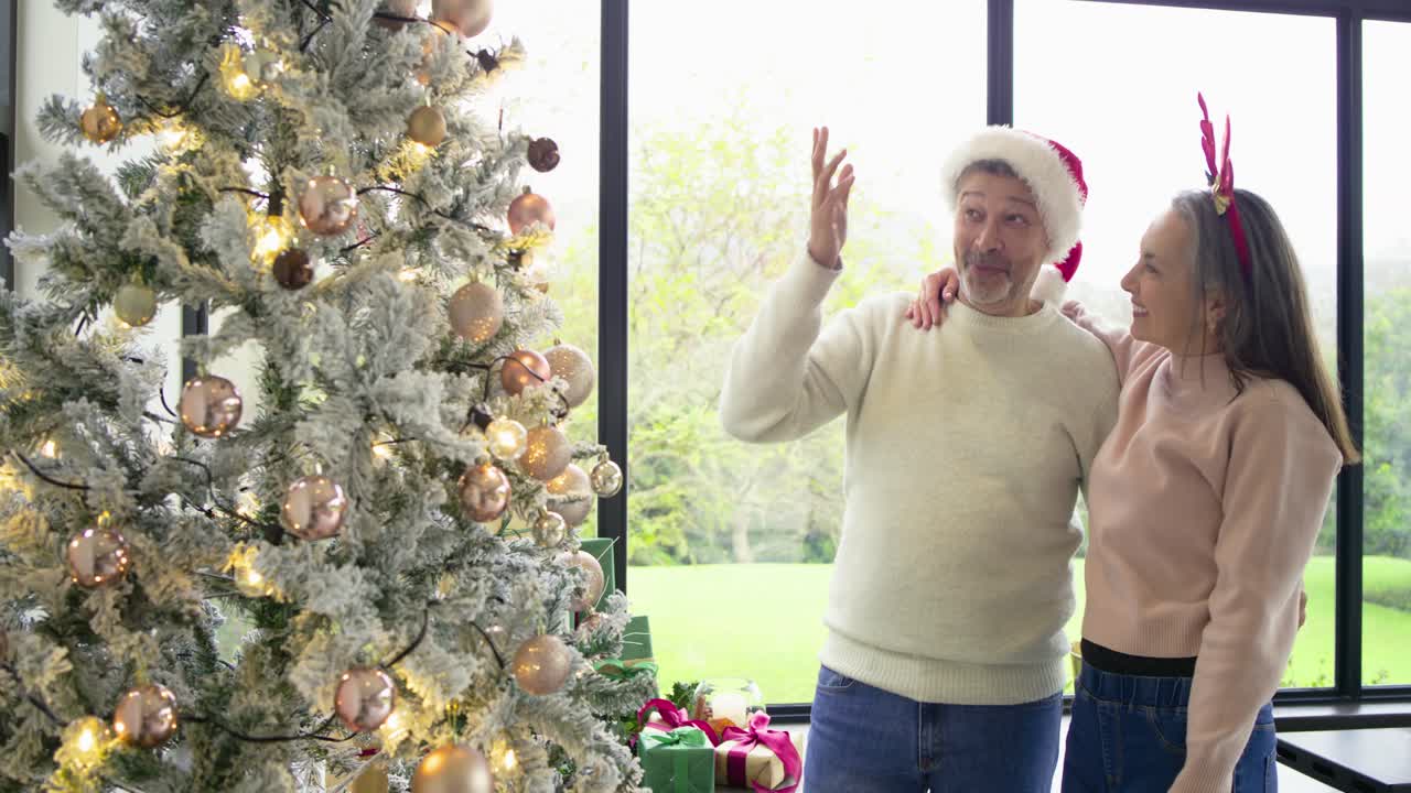 Couple preparing for holiday by decorating Christmas tree with baubles, lights in sunroom embracing