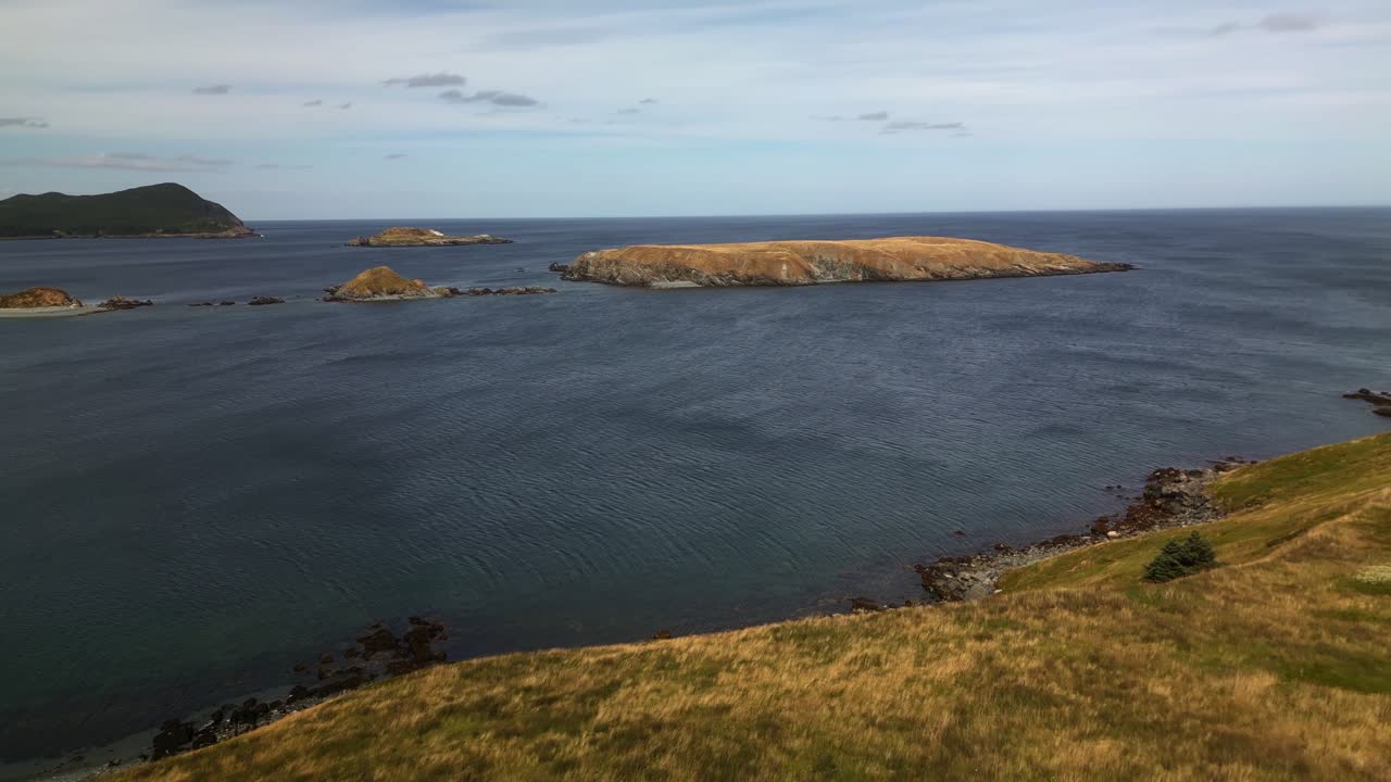 A drone flies over Ferryland’s tranquil bay, revealing calm blue waters dotted with rocky islets and a distant headland, capturing the serene beauty of Newfoundland’s coastline