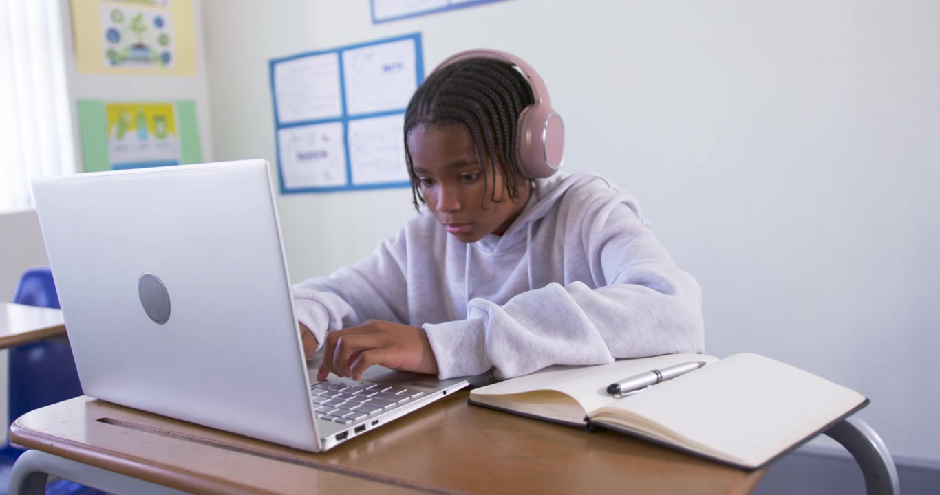 African American teen in gray hoodie, pink headset leaning typing on laptop in class checking notes