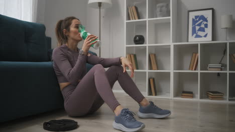 adult woman dressed sportswear is sitting on floor and relaxing after training at home sipping clean water keeping fit and fitness