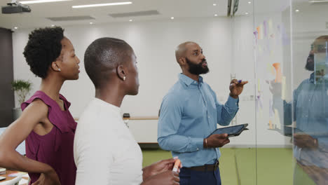 african american colleagues brainstorming, making notes on glass wall in office in slow motion