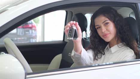 a woman is seated in a land vehicle, holding a car key. the automotive design of the vehicles exterior is visible through the window as she gestures inside the motor vehicle