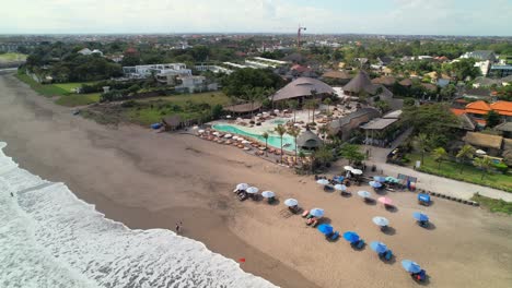 se acerca desde el aire al club de la playa de mari bali desde la playa de batu beleg, el avión no tripulado desciende hacia el área de salón de la piscina rodeada de tumbas y tumbas, olas blancas espumosas rodando sobre la arena suave, indonesia