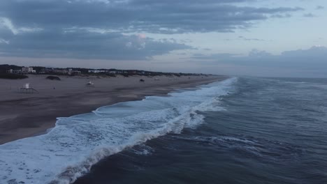 Backwards-flying-shot-of-the-lifeward-stands-on-the-beach-sand-with-water-waves-moving-to-the-sand-in-a-cloudy-day-at-Mar-de-las-Pampas,-Argentina