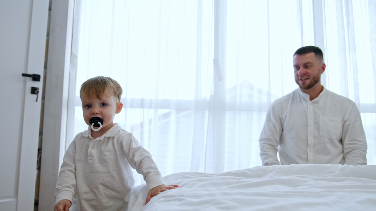 Father and son wearing white t-shirts are in the bedroom. Cute little baby boy run around the bed approaching camera.