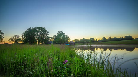 Low-angle-shot-of-sun-rising-over-small-cottages-along-lakeside-in-timelapse-along-lush-green-grasslands