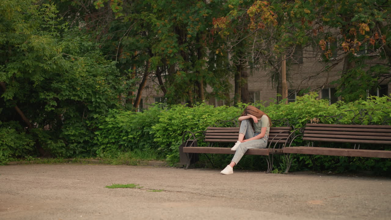 молодая женщина сидит на деревянной скамейке в парке, опираясь головой на руку в отражающей и мрачной позе, в окружении пышной зелени, деревьев и фоновой фигуры, движущейся рядом