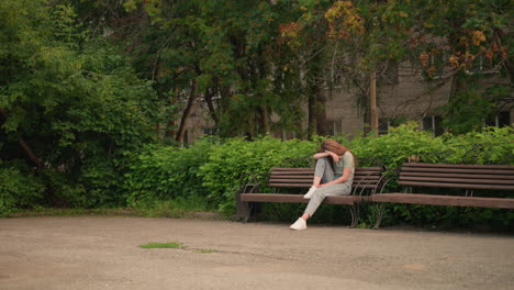 joven sentada en un banco de madera en un parque, apoyando su cabeza en su brazo en una postura reflexiva y sombría, rodeada de exuberante vegetación, árboles y una figura de fondo que se mueve cerca