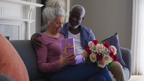 Caucasian-senior-woman-opening-gift-box-while-sitting-with-her-husband-on-the-couch-at-home