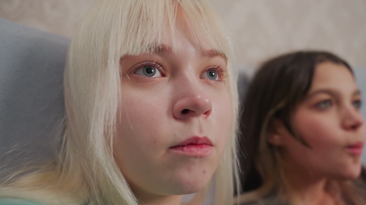 Close up of young blonde girl being fed popcorn by hand while watching movie with friend sitting beside her on bed, both girls appear focused and relaxed in cozy indoor setting with soft lighting