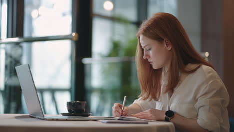 Estudiante-Pelirroja-Está-Aprendiendo-De-Forma-Remota-Haciendo-La-Tarea-En-Un-Libro-De-Ejercicios-Sentado-En-La-Mesa-Con-Una-Computadora-Portátil