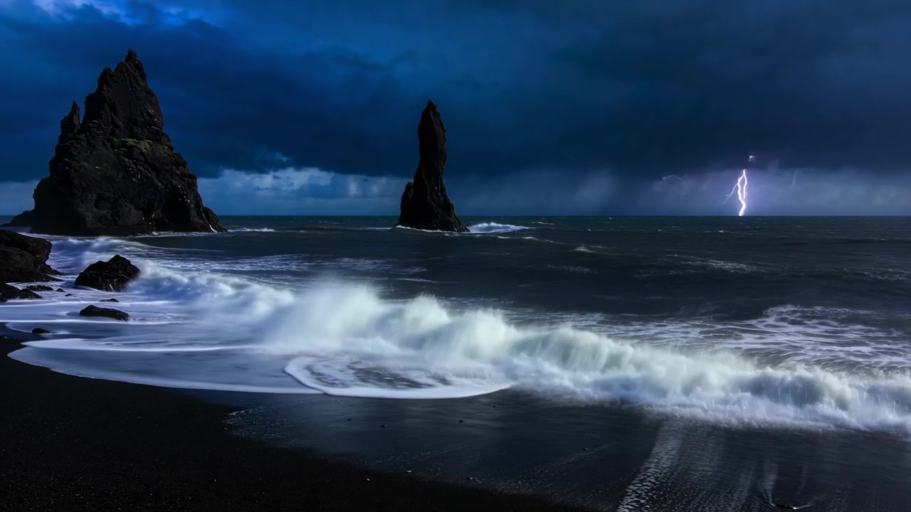 Un oleaje de tormenta entrante empuja olas que rompen sobre una playa de arena negra, golpeando dos farallones en medio de relámpagos.
