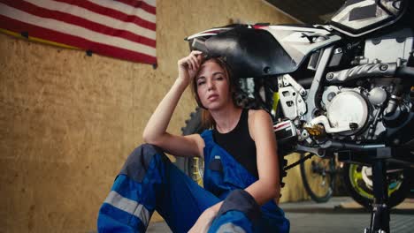 portrait of a tired brunette girl auto mechanic in a black top and blue overalls who folds her hands on her knees leaning on which she has jacked up and is repairing in a workshop with a usa flag on the wall