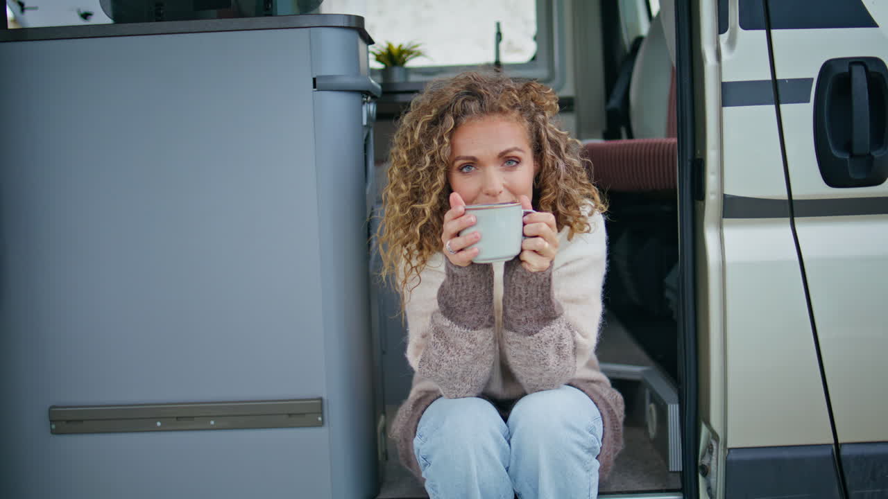 트레일러 계단에 앉아 카푸치노를 마시는 곱슬머리 여자. 행복한 여자가 커피를 즐긴다
