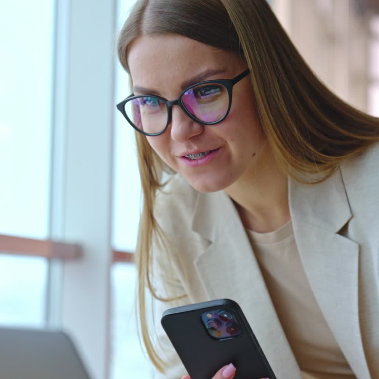 Young woman in glasses and braces holding a phone in hand and looking at laptop. Portrait of a smiling lady working in office. Close up