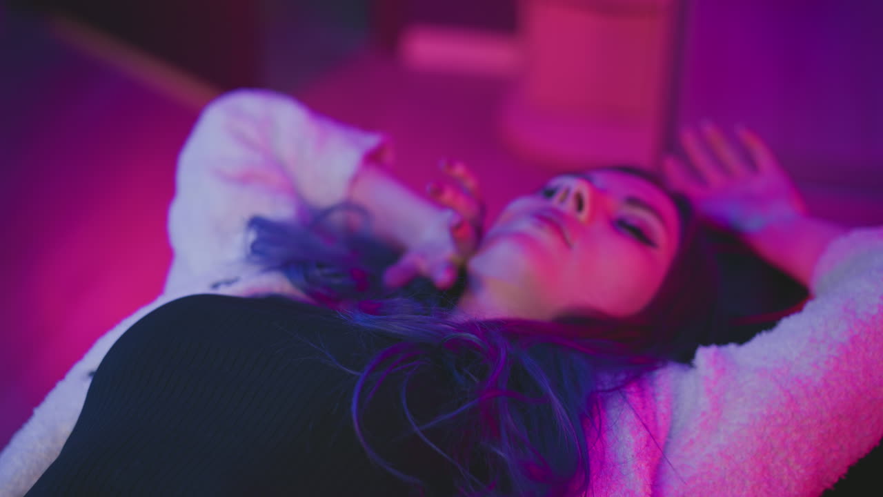 Girl rests peacefully with eyes closed as hand gently touches face in colorful room filled with glowing pink and purple light, moment of sleep in soft cozy attire on reclining chair