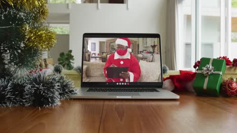 African-american-man-wearing-santa-costume-with-face-mask-on-christmas-video-call-on-laptop
