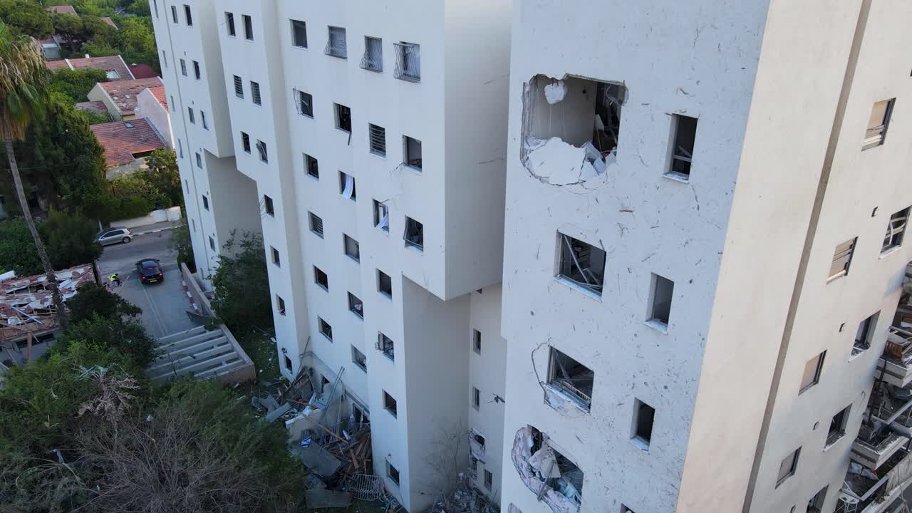 Descending drone shot along a white residential building in Tel Aviv, visibly impacted by a recent strike. A large hole is seen in the structure, with shattered windows, debris on the ground
