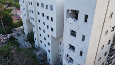 descending drone shot along a white residential building in tel aviv, visibly impacted by a recent strike. a large hole is seen in the structure, with shattered windows, debris on the ground