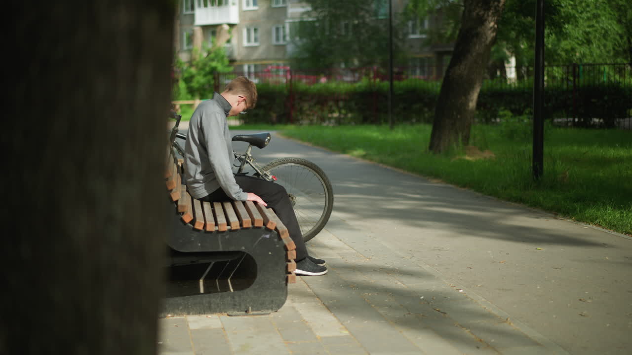 Boy in a gray top and black pants moves away from his parked bicycle, sits down on wooden bench in park, leans back and crosses leg, with a blurry view of a residential building