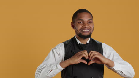 waiter in apron and uniform showing a heart shape sign in studio