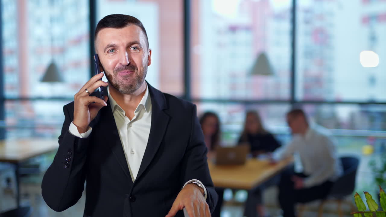 Cheerful positive bearded businessman speaking on the phone in front of camera. Office staff at backdrop in blur.