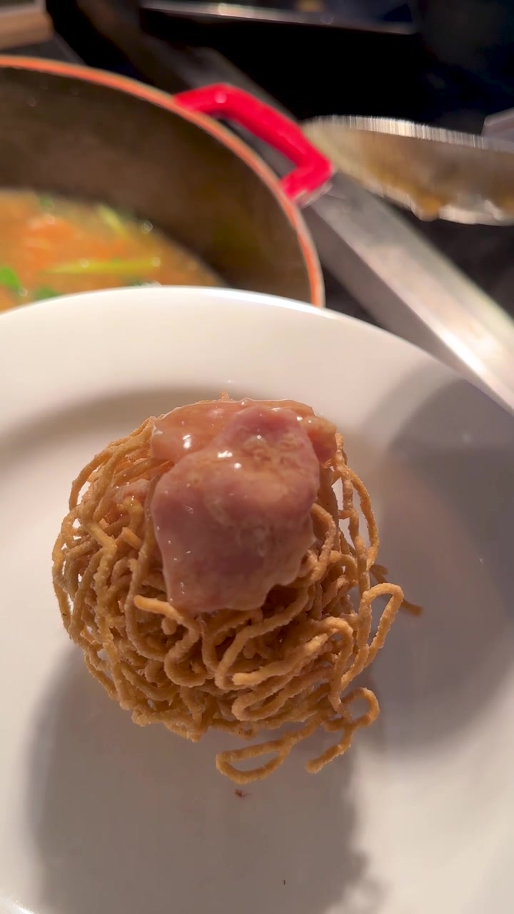 Primo piano di sugo di maiale denso che viene versato con un mestolo su noodles croccanti su un piatto bianco.