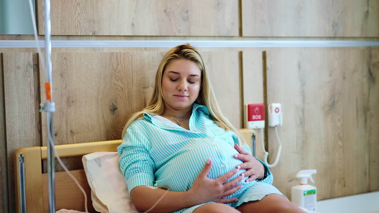 Woman on the last stages of pregnancy lies on bed in hospital. Future mom stroking her big belly while receiving medicine from drop counter.