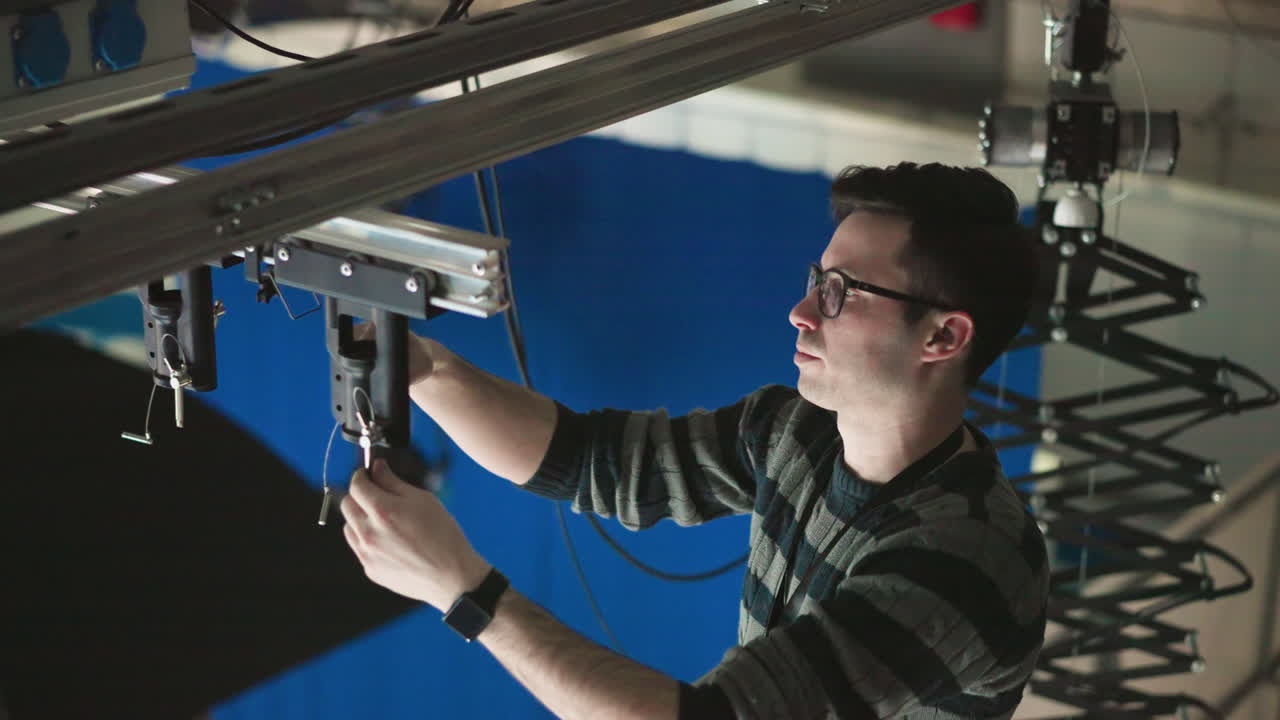 Technician adjusting overhead lighting control unit in studio, focusing on precise positioning and securing equipment, ensuring rigging system is stable and properly aligned for filming or production