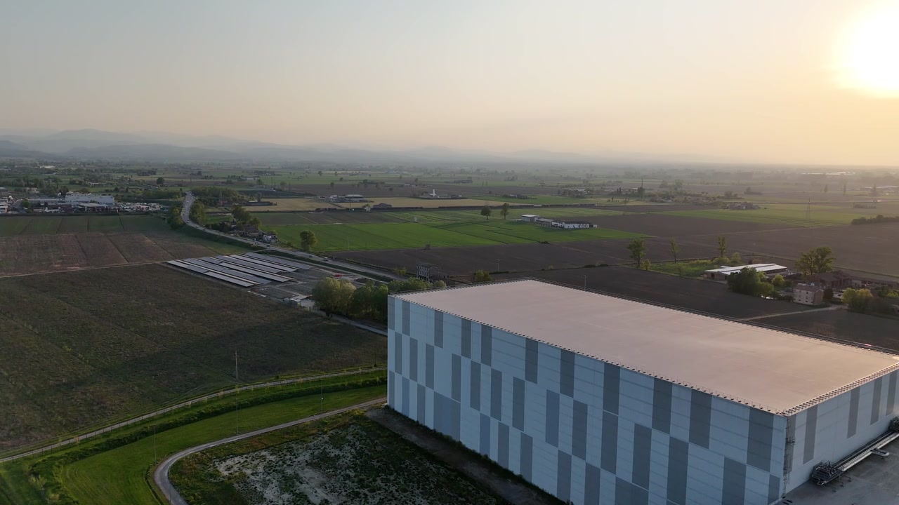 Cold storage logistics facility in Emilia Romagna region, Italy, Europe with large insulated warehouse building beside agricultural fields and solar arrays across Po Valley plain under sunset light