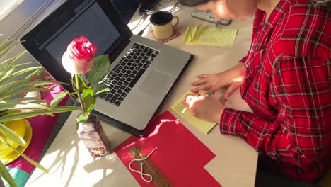 Una-Mujer-Está-Escribiendo-En-Un-Papel-De-Un-Cuaderno-En-El-Escritorio-Mientras-Trabaja-En-Casa-En-Irán,-Teherán,-La-Gente-Iraní-Aprende-El-Idioma-Persa-En-Cursos-En-Línea-Pandémicos,-Clase,-Diseño-De-Decoración-Del-Hogar,-Vestido-Rojo,-Flor-Rosa