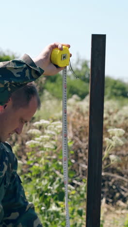 side view skilled repairman in camouflage jacket uses tape for pole height measuring in wild grass on day