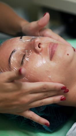 beautician covering female client's face with cellophane. cosmetologist preparing a woman for cosmetic skincare procedures. close up. vertical video