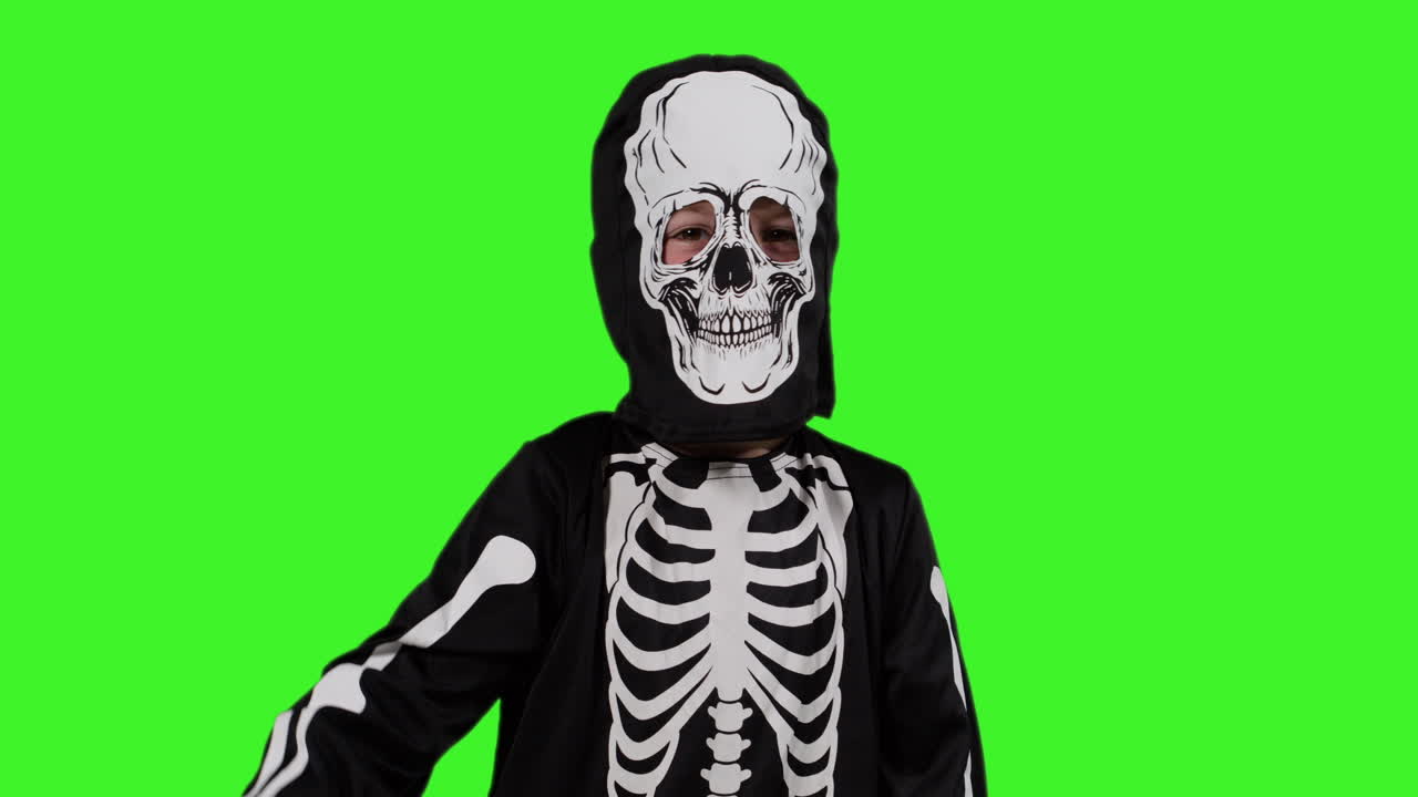 Child Dressed Up In Skeleton Costume Having Fun Trick Or Treating At Halloween Waving At Camera In Front Of Studio Green Screen