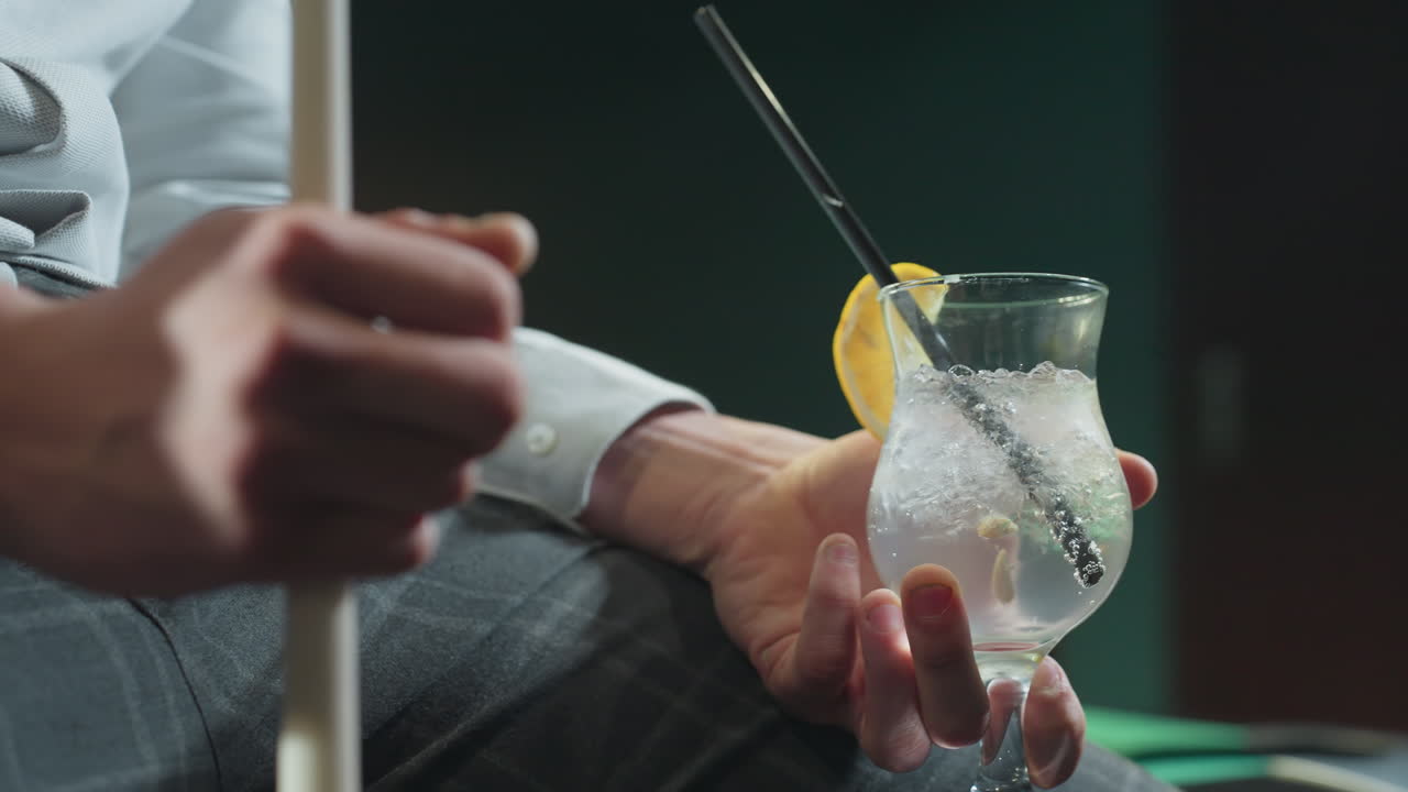 Primo piano di un uomo, volto non visibile, che indossa una camicia bianca e pantaloni grigi, mentre tiene in mano una stecca da biliardo e un bicchiere di limonata con cannuccia nera. Cubetti di ghiaccio e fetta di limone aggiungono dettagli rinfrescanti.