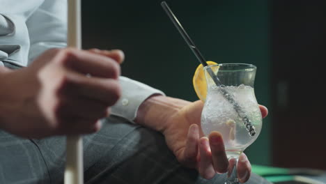 close-up of guy, face not visible, wearing white shirt and grey trousers, holding cue stick and glass of lemon drink with black straw. ice cubes and lemon slice add refreshing details