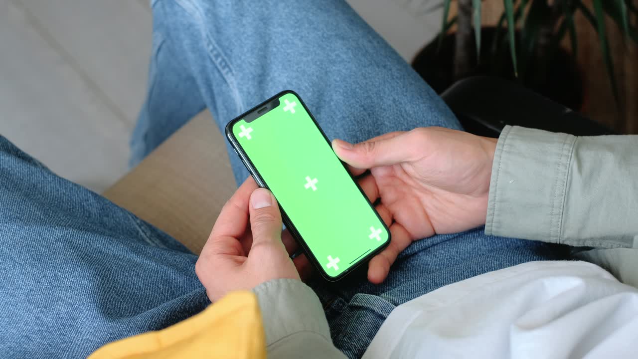 Uomo seduto in poltrona a casa usa lo smartphone con mockup schermo verde. 4K