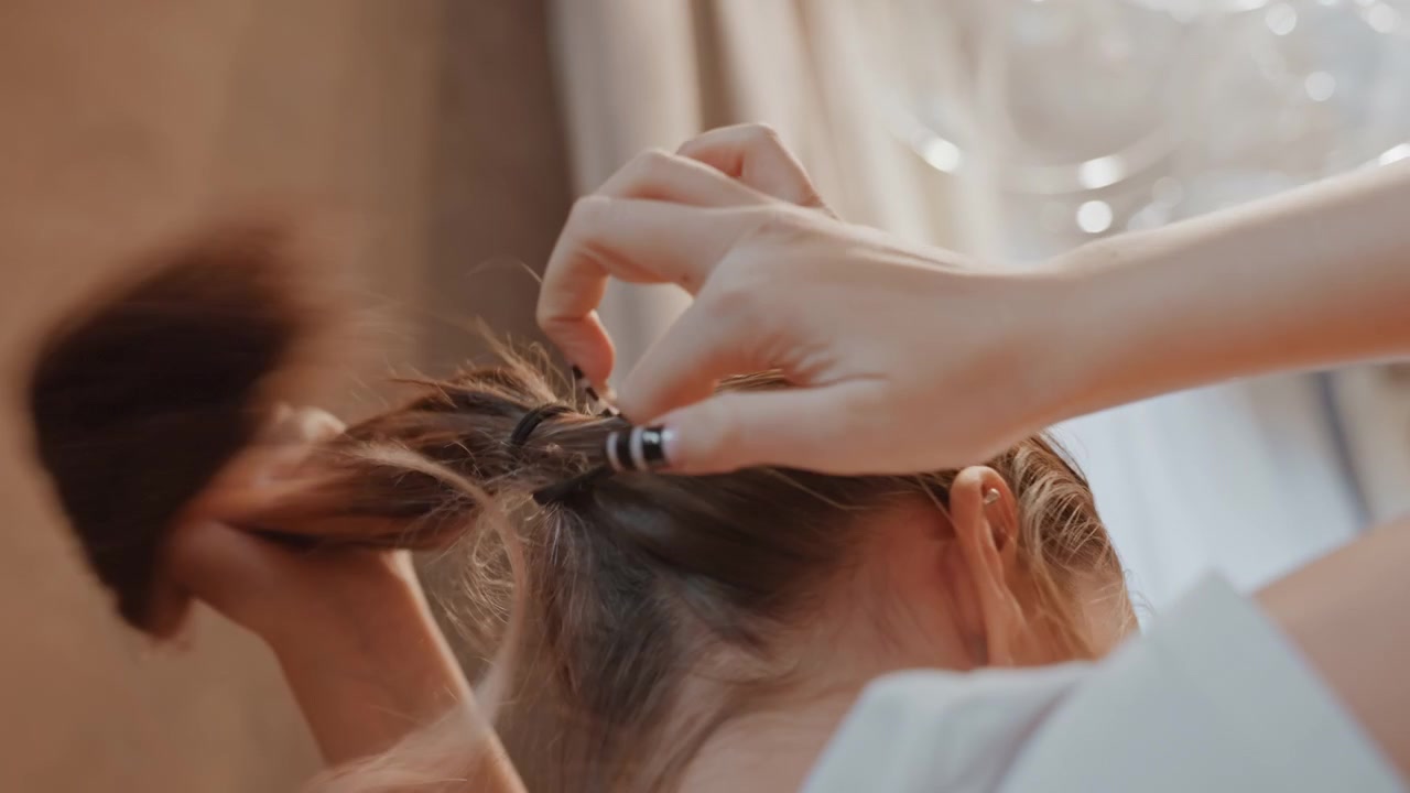 Bedroom scene of woman grooming. Casual woman arranging ponytail near window. Closeup of woman tying her hair in cozy bedroom. Gentle moment of woman securing her ponytail by soft bedroom light