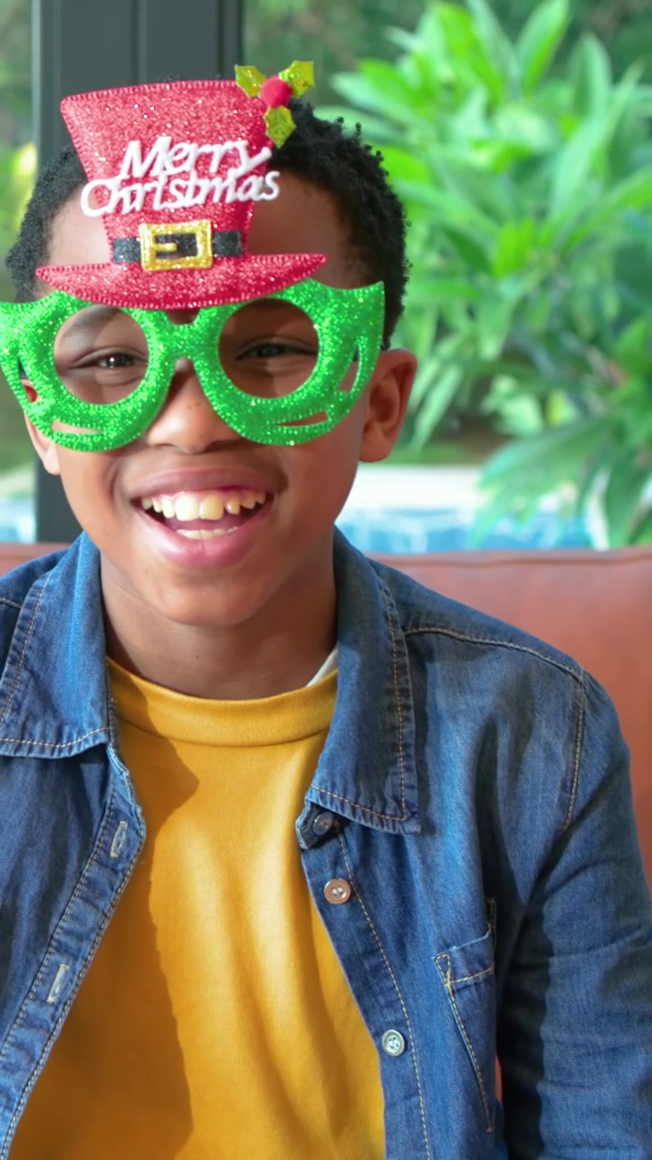Vertical video: Bringing green glasses, boy wearing yellow shirt on sofa placing prop for Christmas