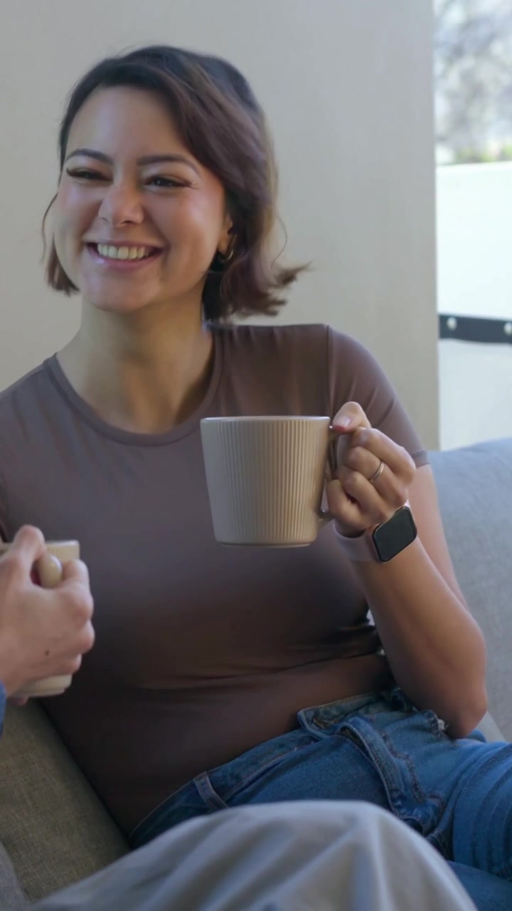 Video vertical: Mujer con camiseta marrón y taza, con reloj en el sofá, respondiendo al comentario de una amiga