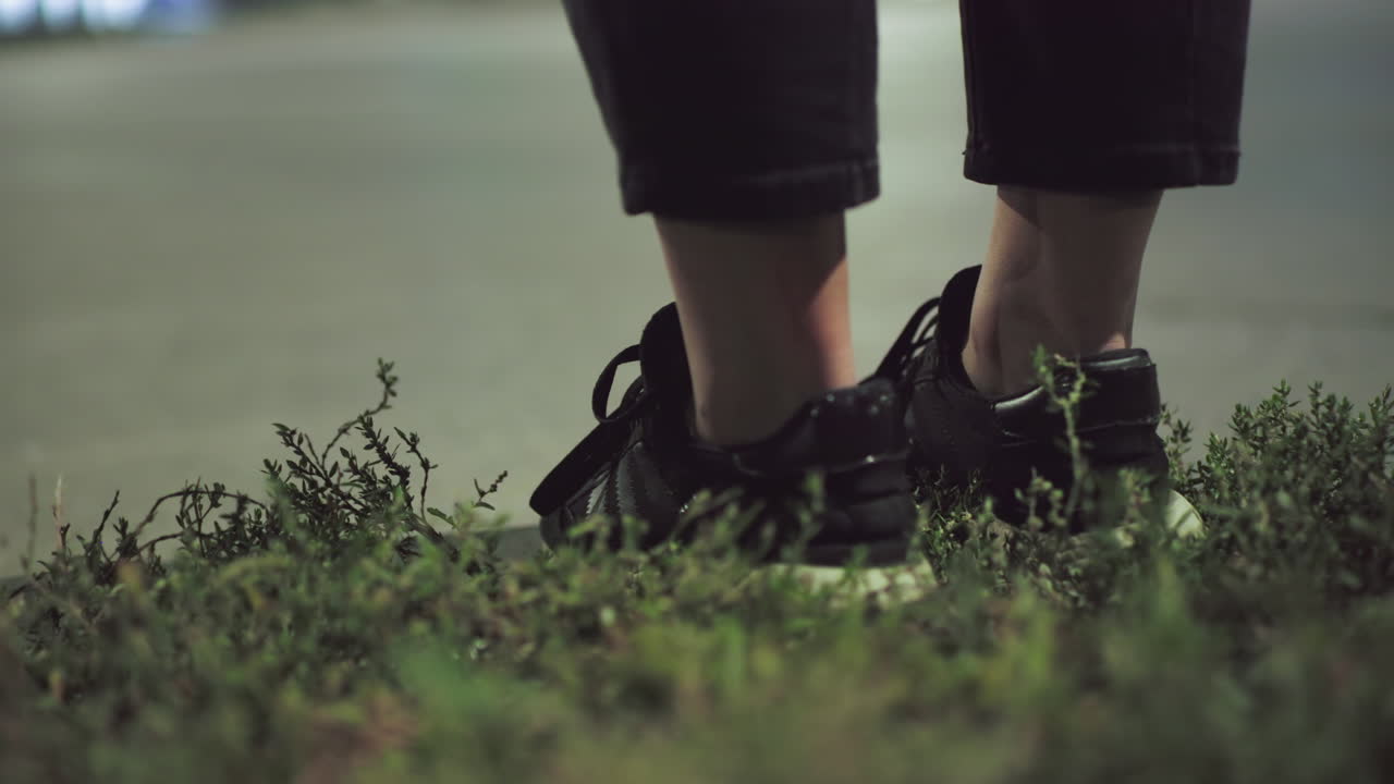 Leg view of individual in black sneakers standing by roadside at night with grass in foreground and blurred passing traffic in background illuminated by soft ambient streetlight
