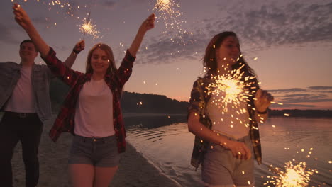 Alegres-Amigos,-Hombres-Y-Mujeres,-Corren-Por-La-Playa-Al-Atardecer-Sosteniendo-Brillantes-Fuegos-Artificiales-Y-Luces-Desbocadas-En-Cámara-Lenta.-Baile-Y-Fiesta-Al-Atardecer-En-La-Playa