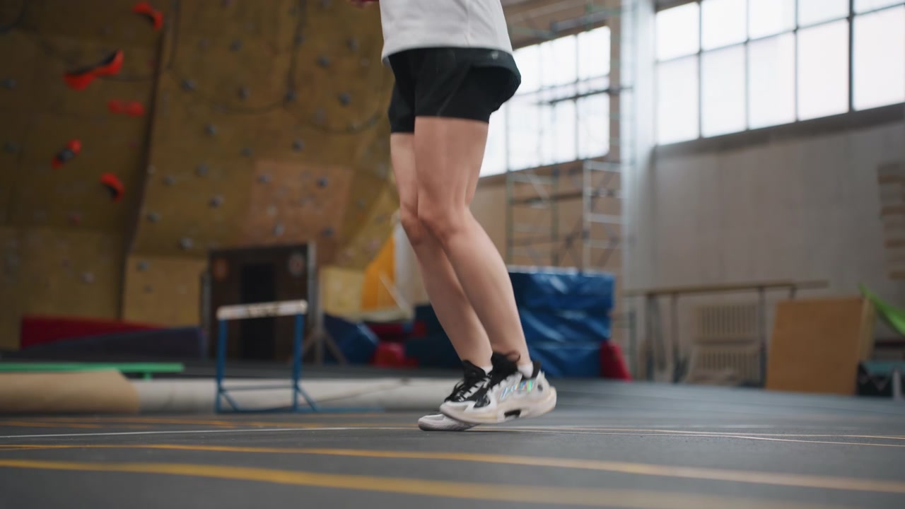 Indoor-Sporthalle: Weißer Athlet übt Badminton-Beinarbeit, Nahaufnahme von Beinen und Turnschuhen, schnelle Sprünge und präzise Landungen, intensive Trainingsatmosphäre, Trainerpräsenz und Kletterwand als Hintergrund, fokussierte Agilität