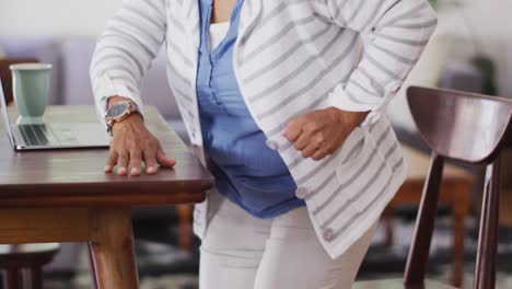 Eine-Asiatische-ältere-Frau-Hält-Sich-Vor-Schmerzen-Im-Rücken,-Während-Sie-Zu-Hause-Von-Einem-Stuhl-Aufsteht