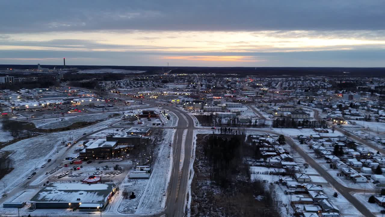 Luchtfoto van Thompson, Manitoba in november met sneeuwbedekte grond. De drone vliegt over de snelweg richting de stad langs huizen, lichten en een gedeeltelijk bewolkte schemerlucht