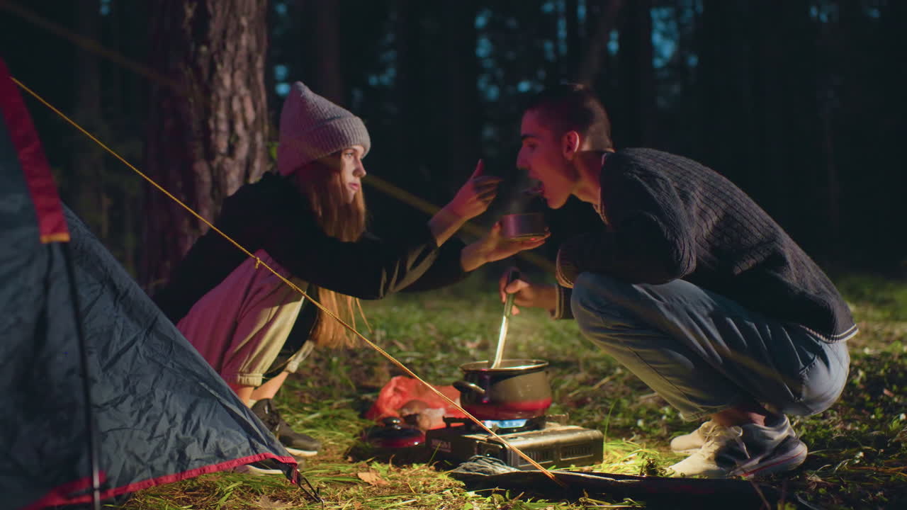 Woman feeds man with canned food during night camping near forest as they sit beside tent and cook over portable stove, surrounded by trees and soft light