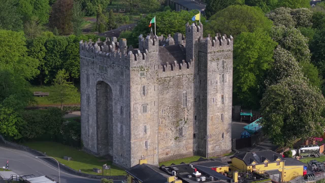 Bunratty Castle In County Clare, Ireland - Stone Fortress With Battlements And Irish Flags Surrounded By Lush Greenery. static aerial shot
