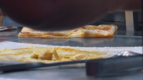 Toma-Cercana-De-Agregar-Rodajas-De-Plátano-A-Crepes-En-El-Mercado-Nocturno-Para-Comida-Callejera