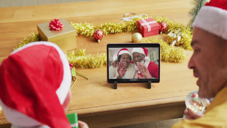 Padre-E-Hijo-Caucásicos-Con-Gorros-De-Papá-Noel-Usando-Una-Tableta-Para-Una-Videollamada-Navideña-Con-Una-Pareja-En-La-Pantalla