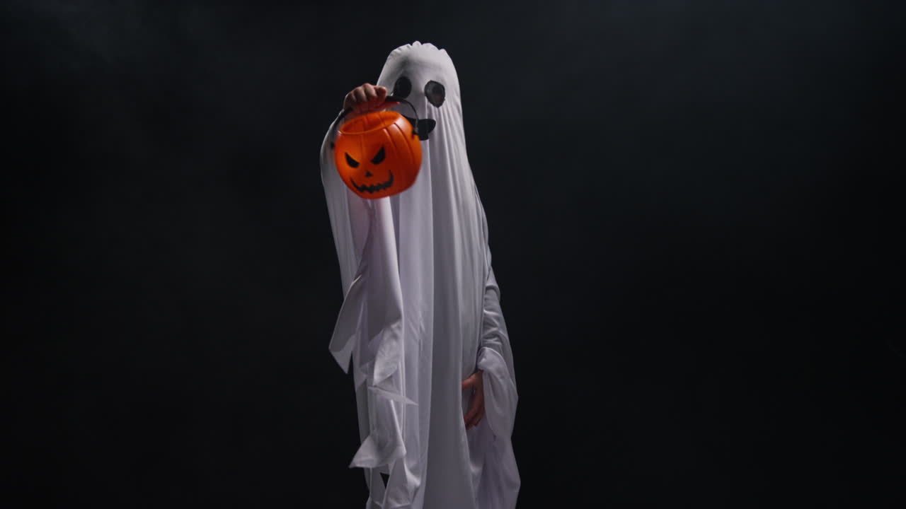 niño vestido con disfraces de fantasma truco o trato en halloween sosteniendo balde en forma de calabaza jack o'lantern contra fondo negro